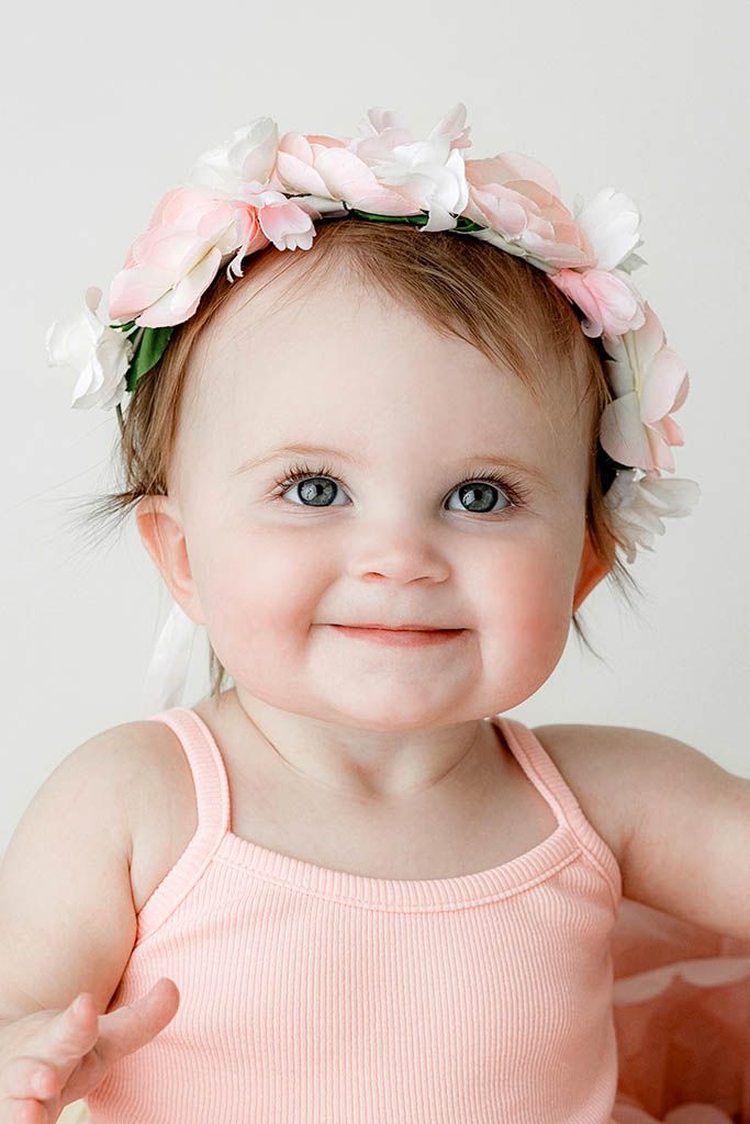 Adorable baby girl with blue eyes wearing a pink floral crown headband and pink outfit, smiling sweetly at the camera.