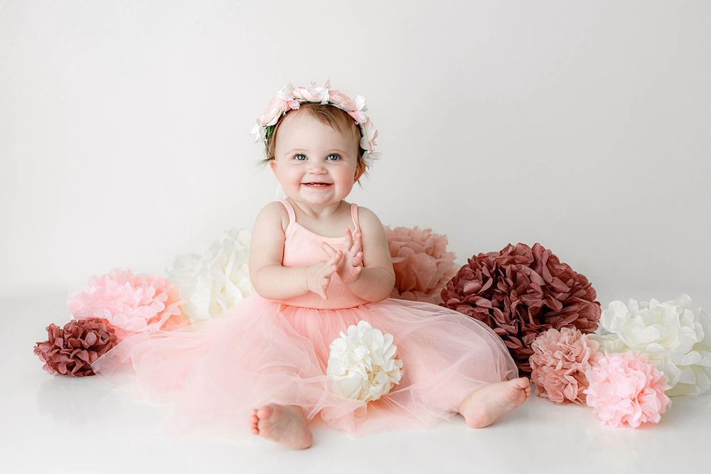 Smiling baby girl in pink tutu dress and floral crown surrounded by colorful paper pom poms on white background.