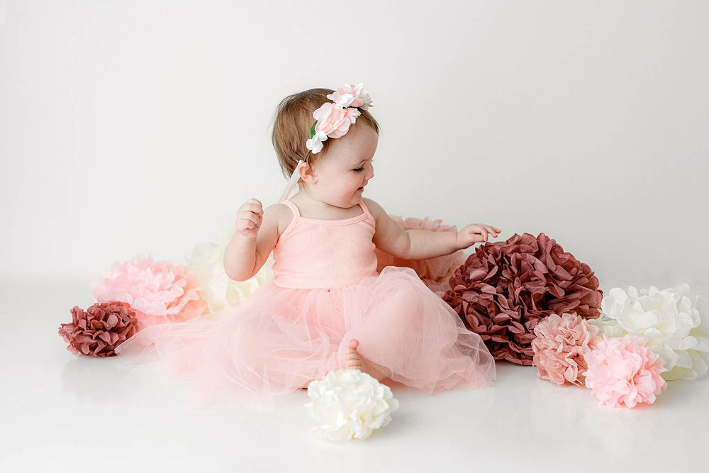 Adorable baby girl in pink tulle dress with floral headband playing among decorative paper flowers on white backdrop.