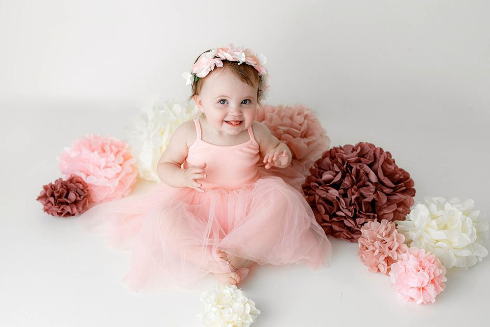 Happy baby girl in a pink tutu dress sitting among decorative paper flowers during a milestone photo session.
