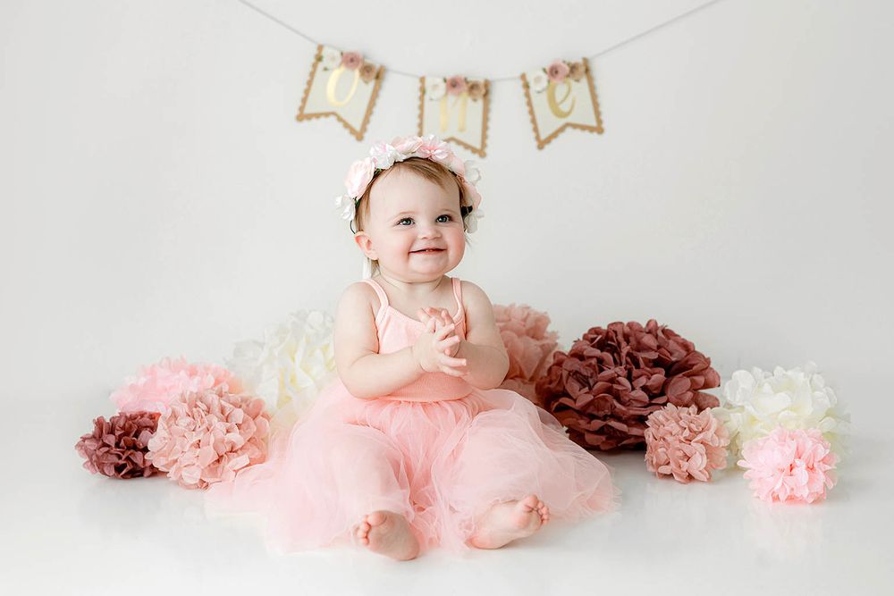 Baby girl in pink tutu sits on floor smiling near paper flowers with ONE birthday banner.