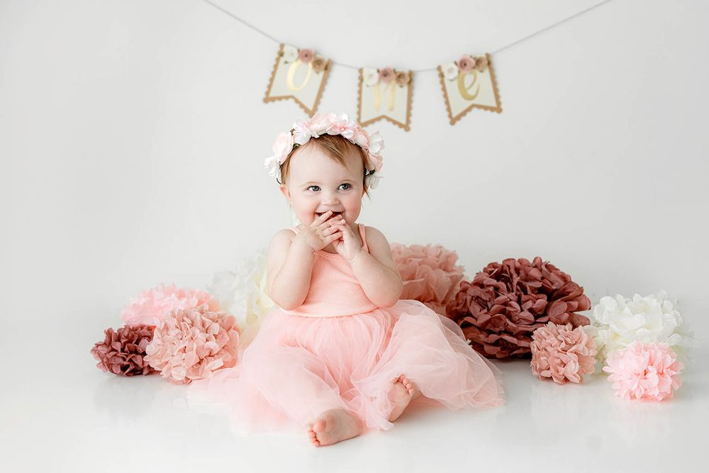 Cute baby in pink tutu sitting on floor with fingers in mouth near colorful paper flowers.