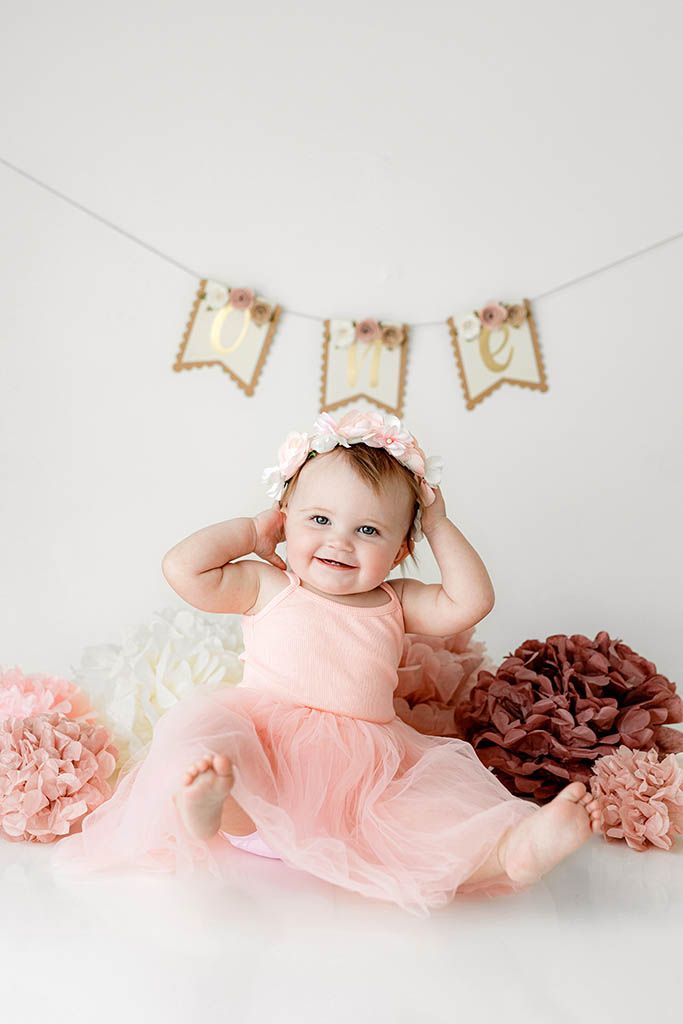 Smiling baby girl in pink tutu dress poses at one year cake smash session with floral banner and pom poms.