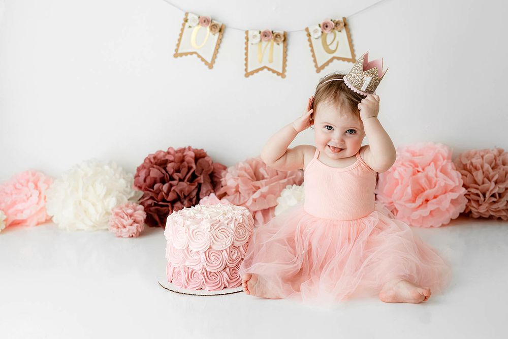 Baby girl in pink tutu dress wearing gold crown poses beside pink rosette cake at first birthday cake smash session.