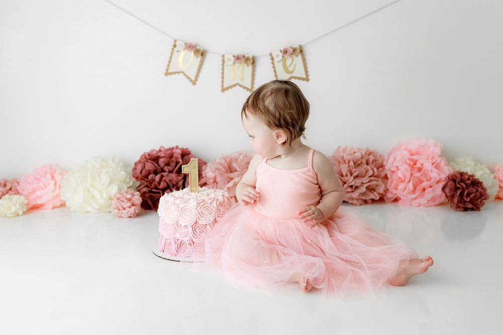 Baby girl in pink tutu dress sits with smash cake at first birthday photo session with floral decorations.