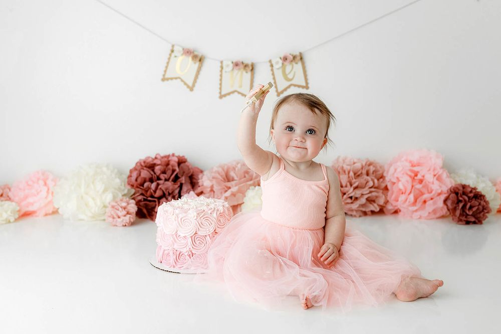 Adorable baby girl in pink tulle dress reaches up beside smash cake at first birthday photo session.