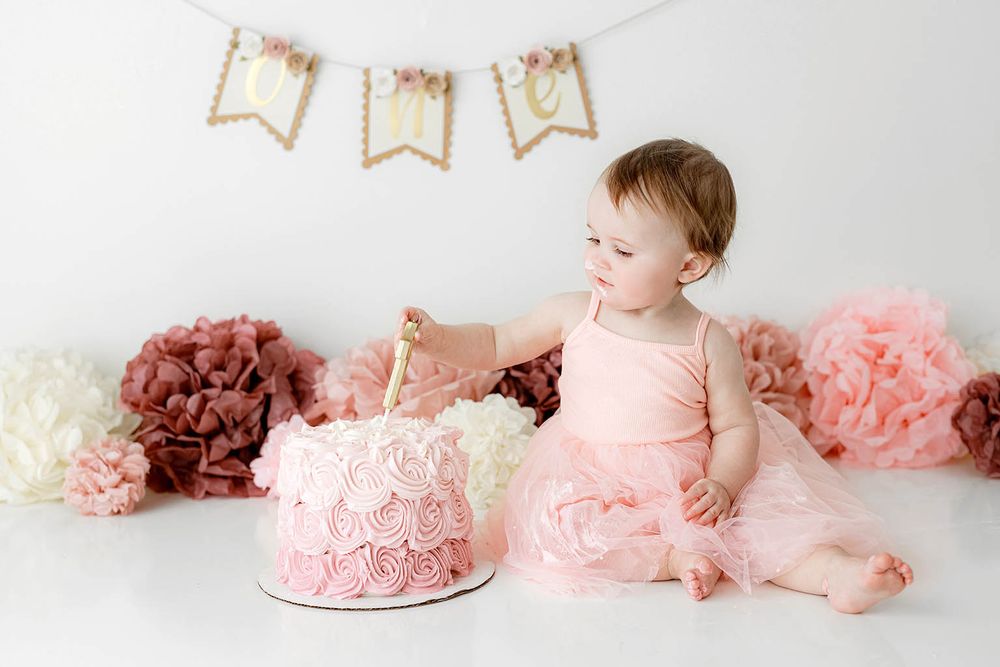 Baby girl in pink tutu smashing first birthday cake surrounded by paper flowers and ONE banner.