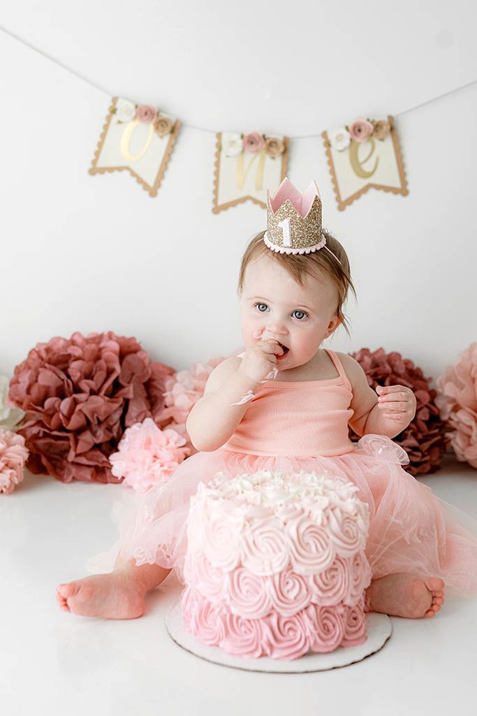Adorable baby girl in pink tutu tastes first birthday cake during cake smash photo session with gold ONE banner.