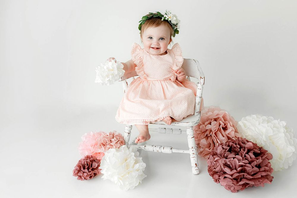 Baby girl in pink dress and floral crown sits on white chair surrounded by decorative paper flowers.