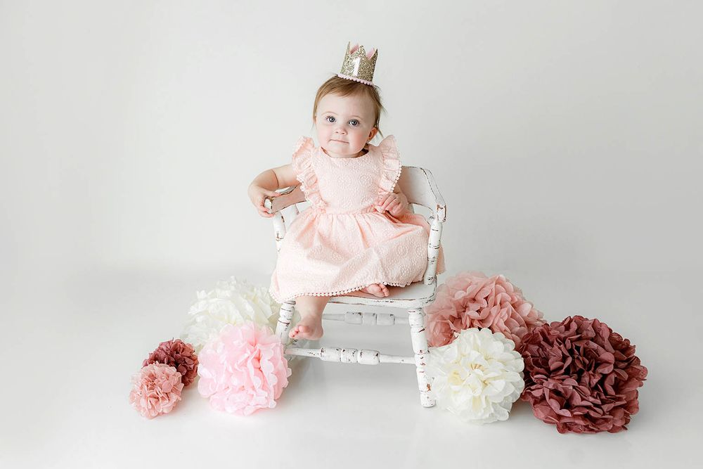 Baby girl in pink dress and crown sits on white chair surrounded by pink and mauve paper flowers.