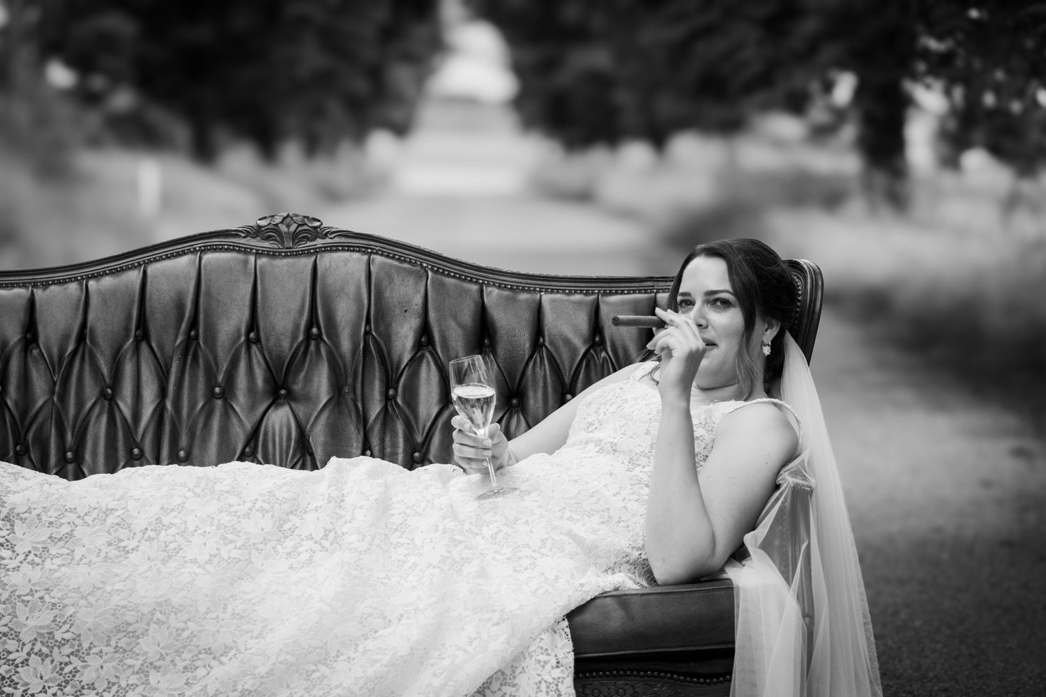 Elegant bride in lace gown reclines on vintage sofa holding champagne in black and white photo.
