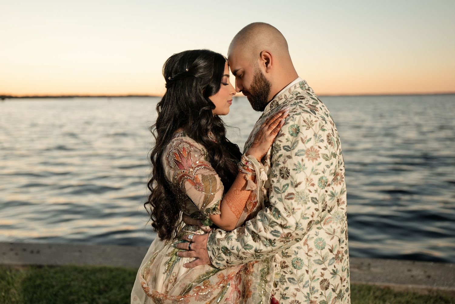 A couple in ornate South Asian attire poses by a waterfront at sunset, the man in embroidered sherwani, woman in floral lehenga.