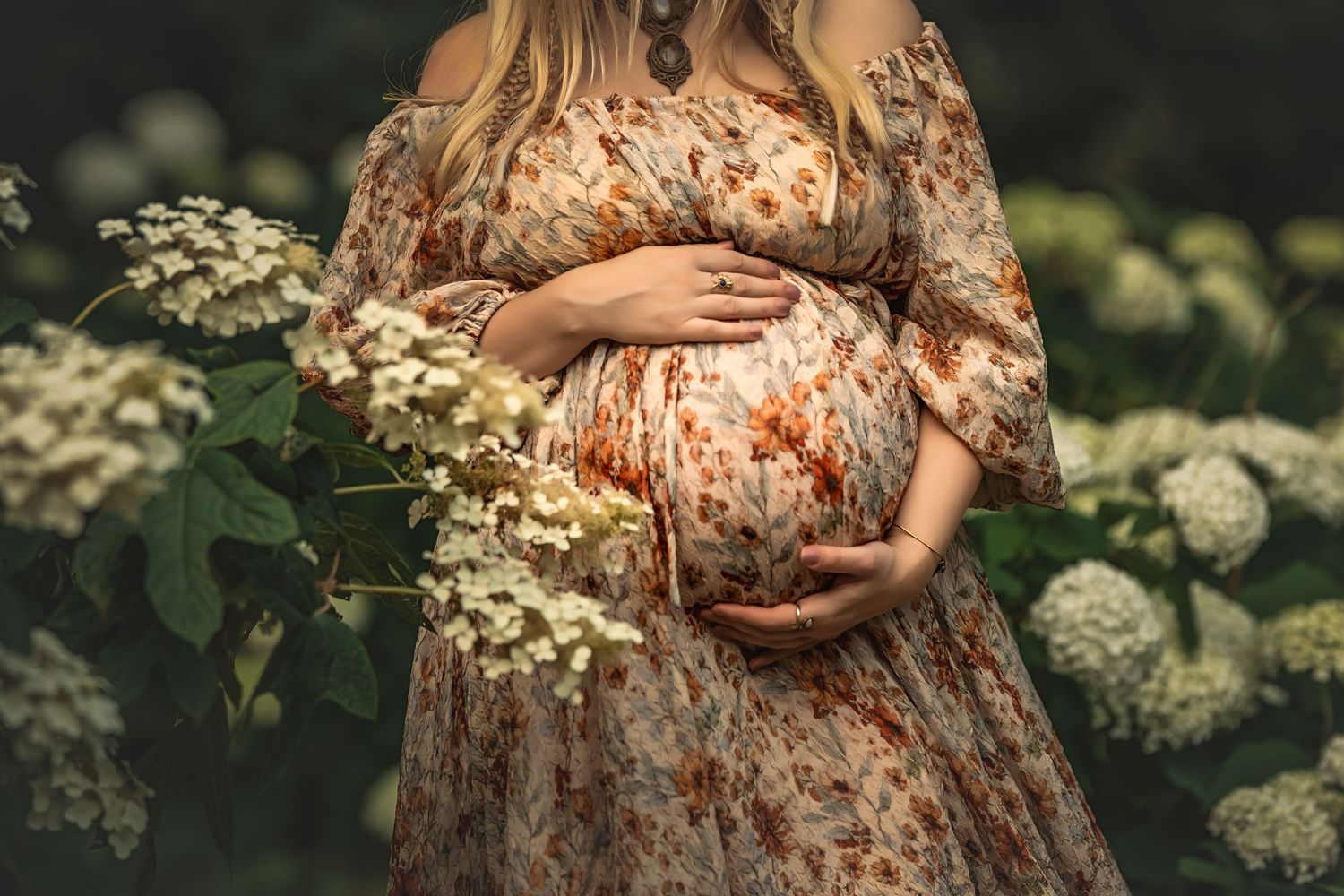 Pregnant woman in floral dress cradling baby bump surrounded by white hydrangea flowers in lush garden by Atlanta maternity photographer.