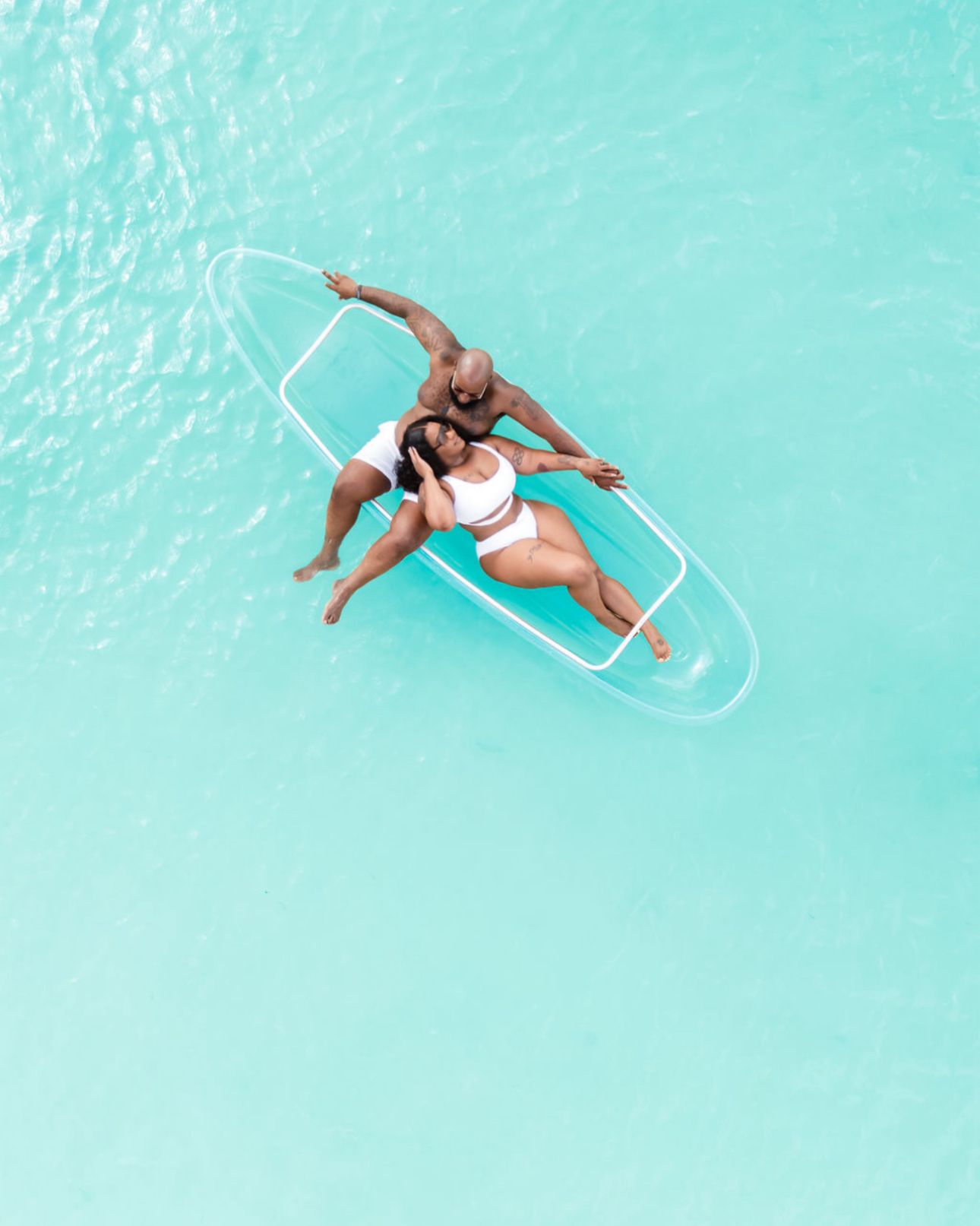 Aerial drone view of couple relaxing together on transparent clear paddleboard in turquoise tropical ocean.