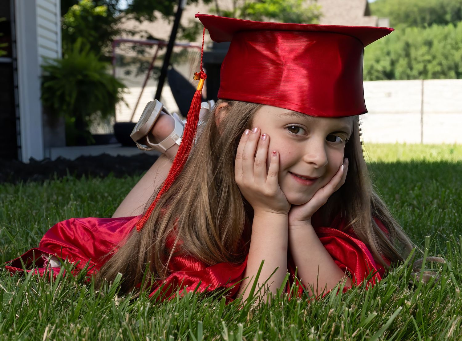 Young girl in red graduation cap and gown lying on grass, smiling with hands on cheeks.