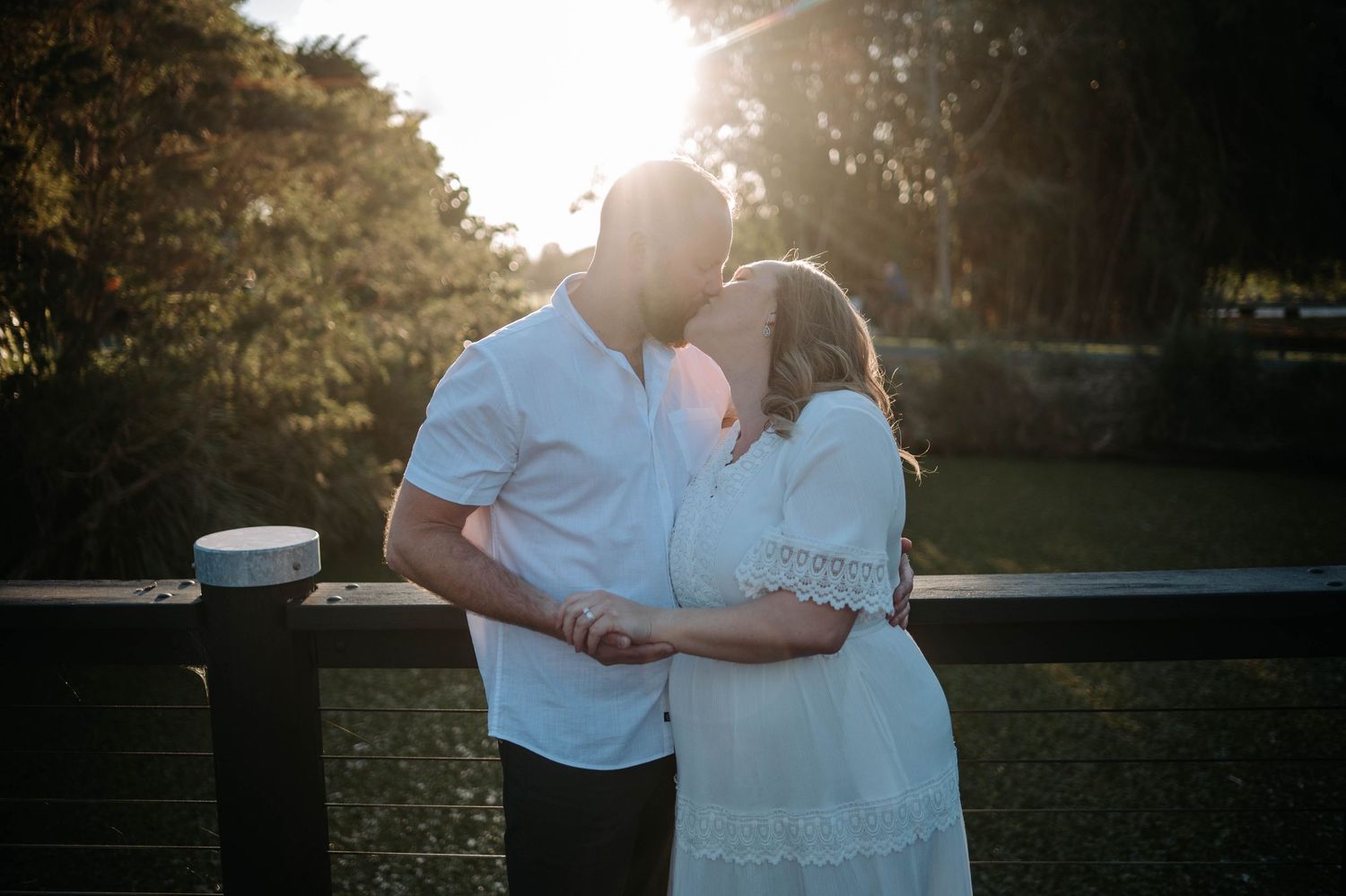 Couple sharing a romantic moment at sunset, golden backlight glowing behind them in a lush green outdoor setting.