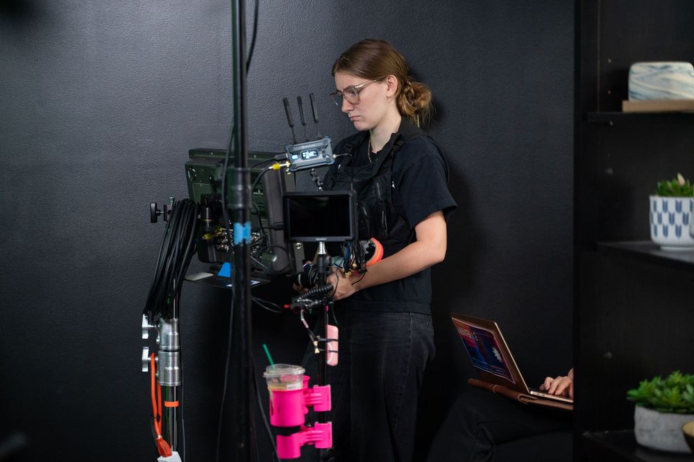 A young woman operating a professional film camera rig in a dark studio setting.