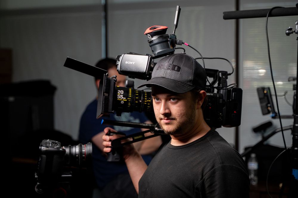 A cameraman holds a professional video camera on his shoulder while filming on a dimly lit production set.