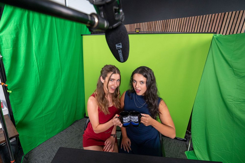 Two women pose together in front of a green screen in a professional video production studio setting.