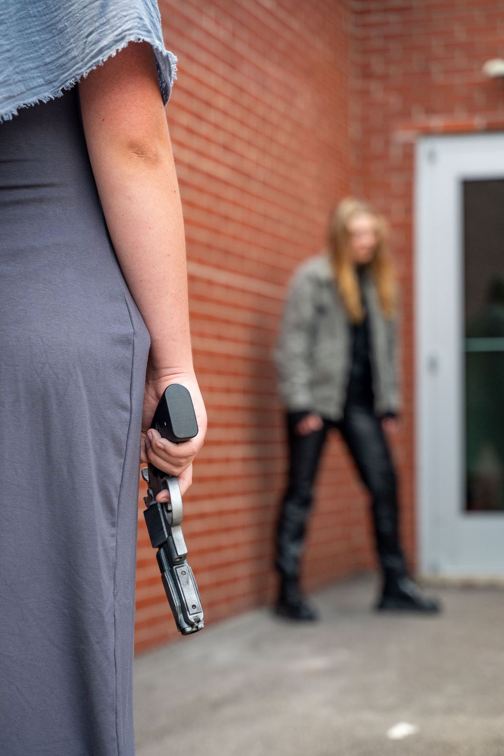 Woman concealing a handgun behind her back while another woman stands in the background near a brick wall.