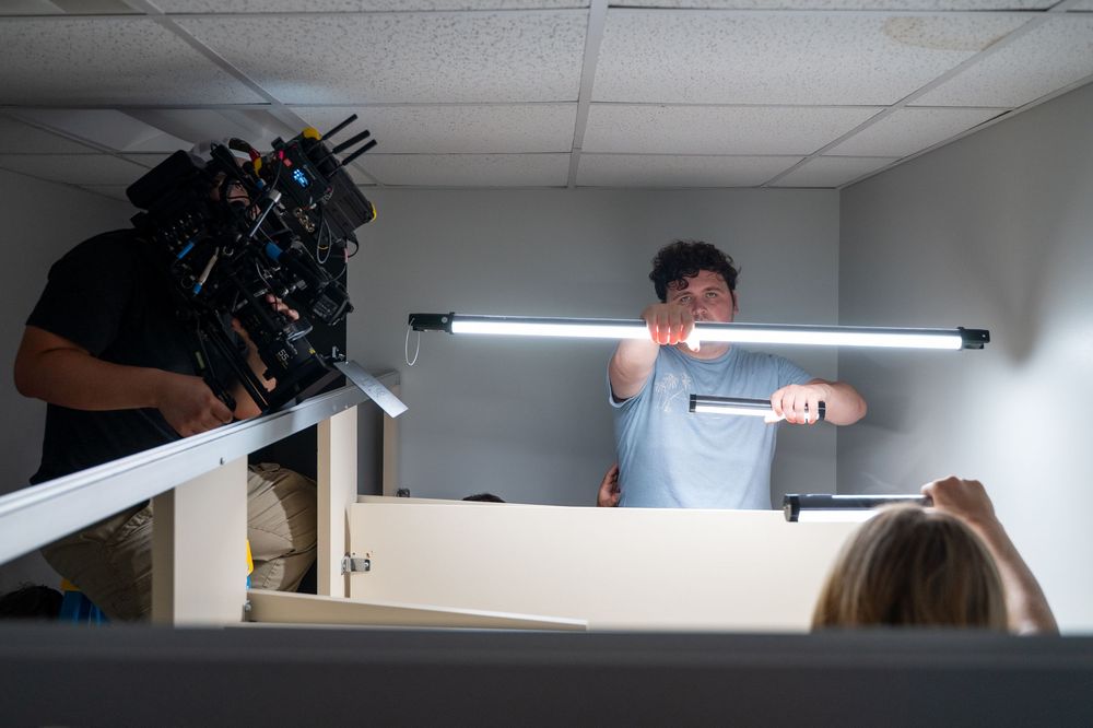 Film crew member holds fluorescent light tube on an indoor set while a cameraman films the scene.