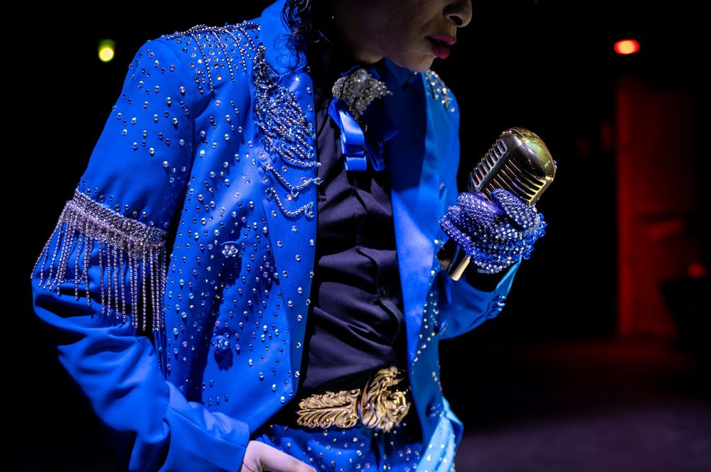 Performer in a blue rhinestone-studded jacket with gold trim holds a vintage microphone on stage.