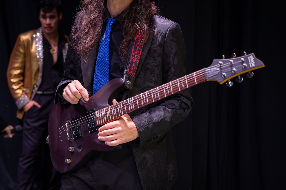 Musician in black suit playing purple electric guitar on stage with performer in gold jacket behind.