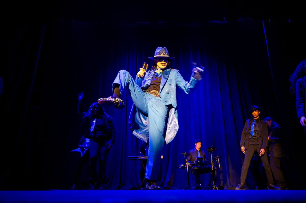 Performer in blue suit and hat dancing on stage under dramatic blue lighting during a theatrical production.