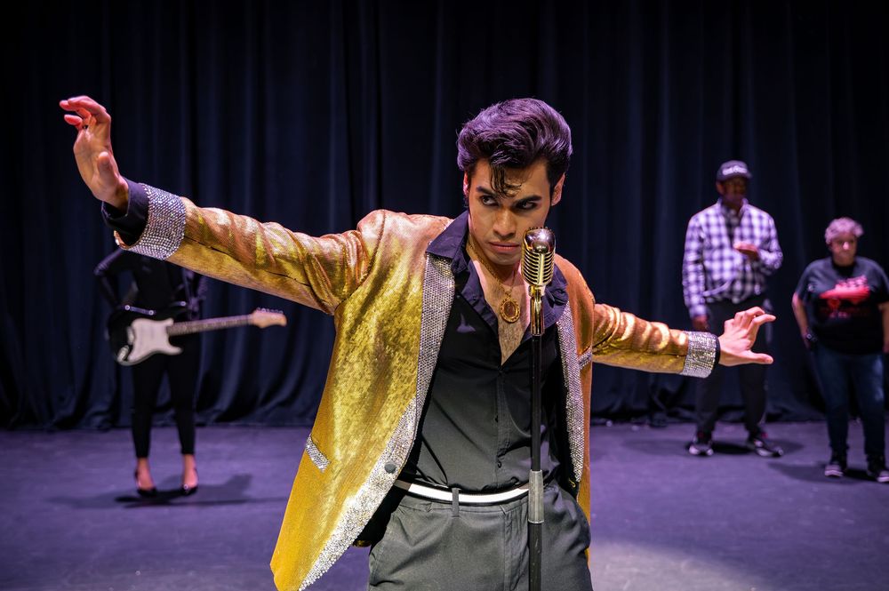 Performer in gold sequin jacket dances on stage with arms outstretched, backed by musicians in a darkened theater.