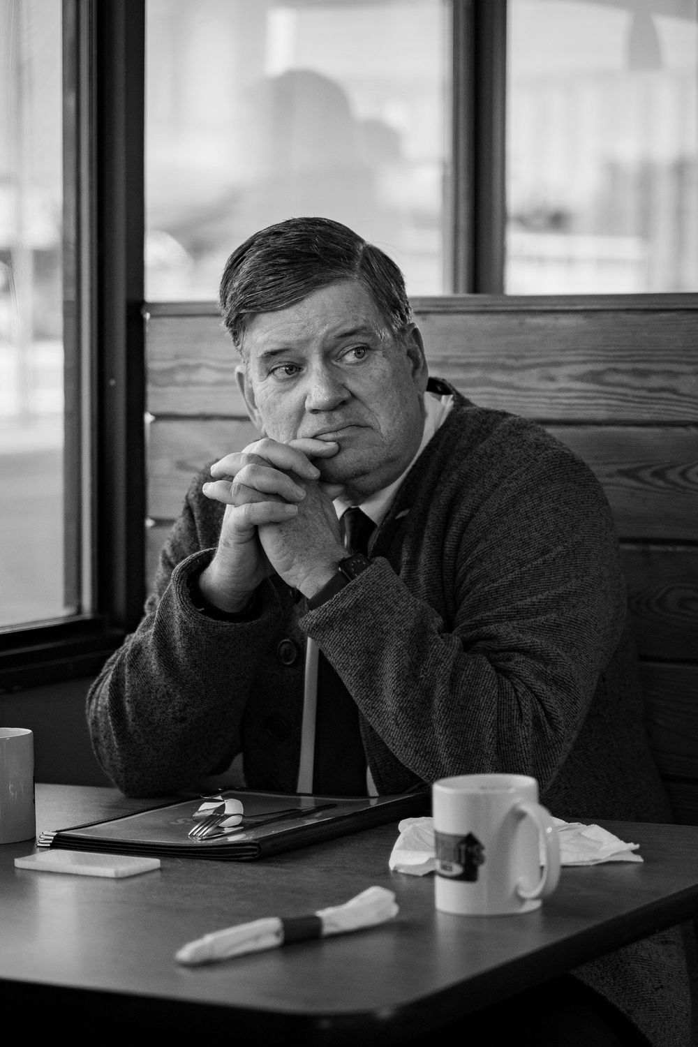 Black and white portrait of a middle-aged man in a suit sitting pensively at a diner table with coffee mugs.