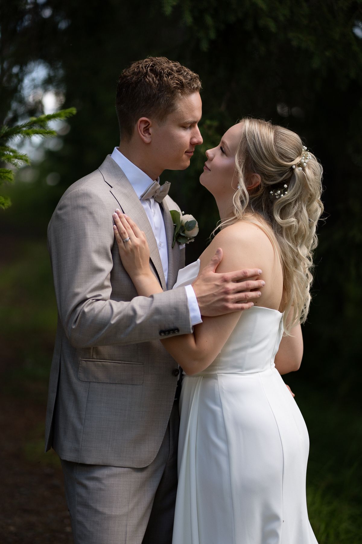 A bride and groom share a romantic almost-kiss moment outdoors, dressed in white gown and gray suit with boutonniere.