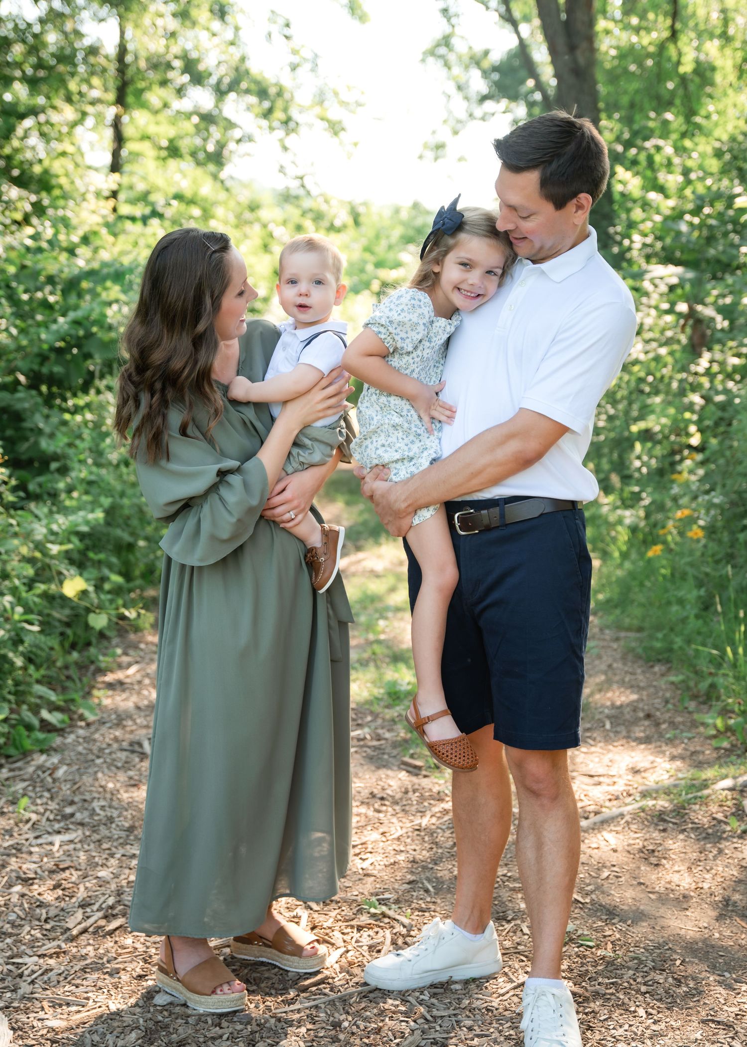 A happy family of four poses on a wooded path, parents holding two young children in coordinated green and white outfits.
