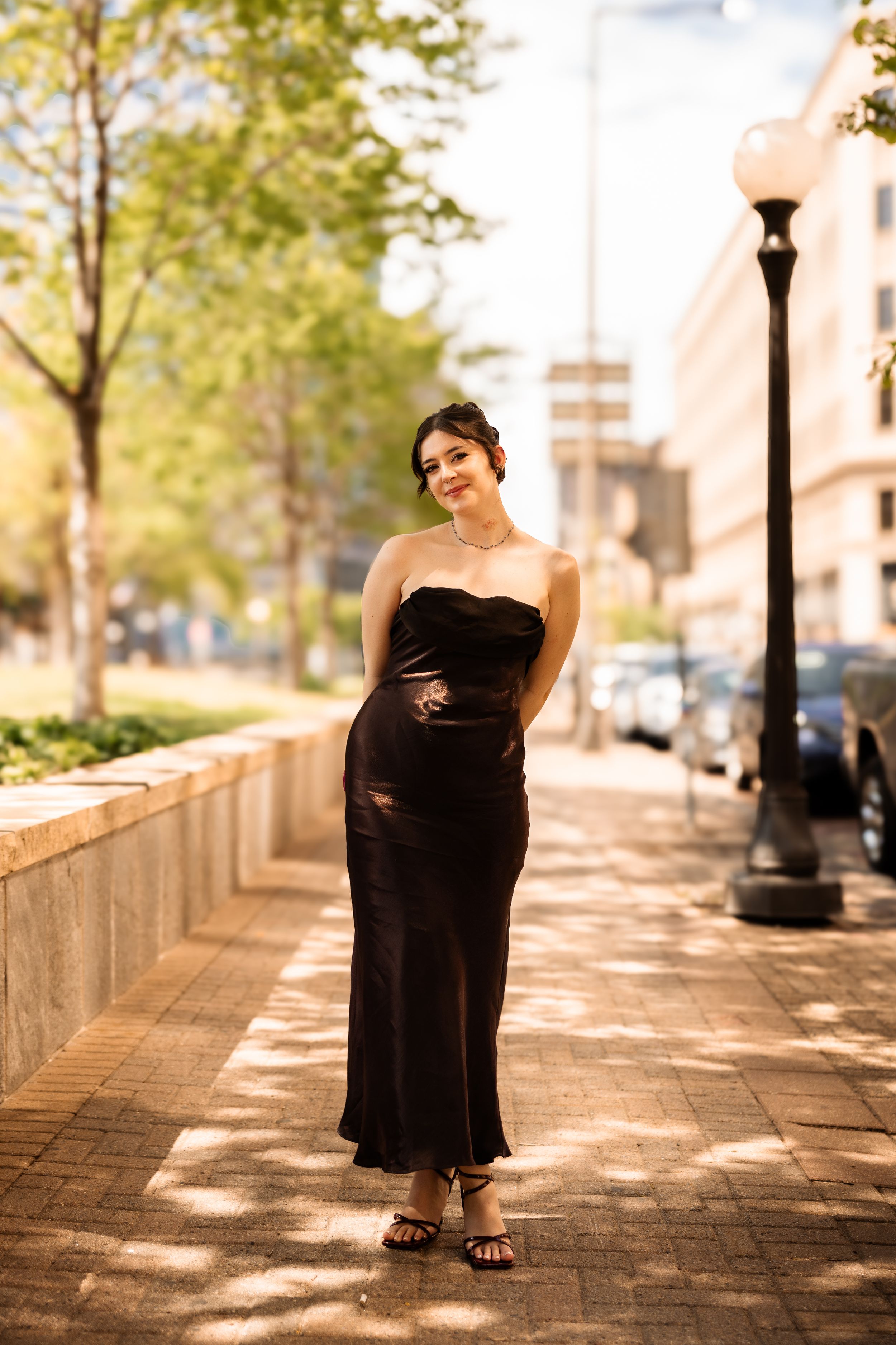 Woman in elegant black strapless midi dress posing on urban brick sidewalk with trees and street lamp.