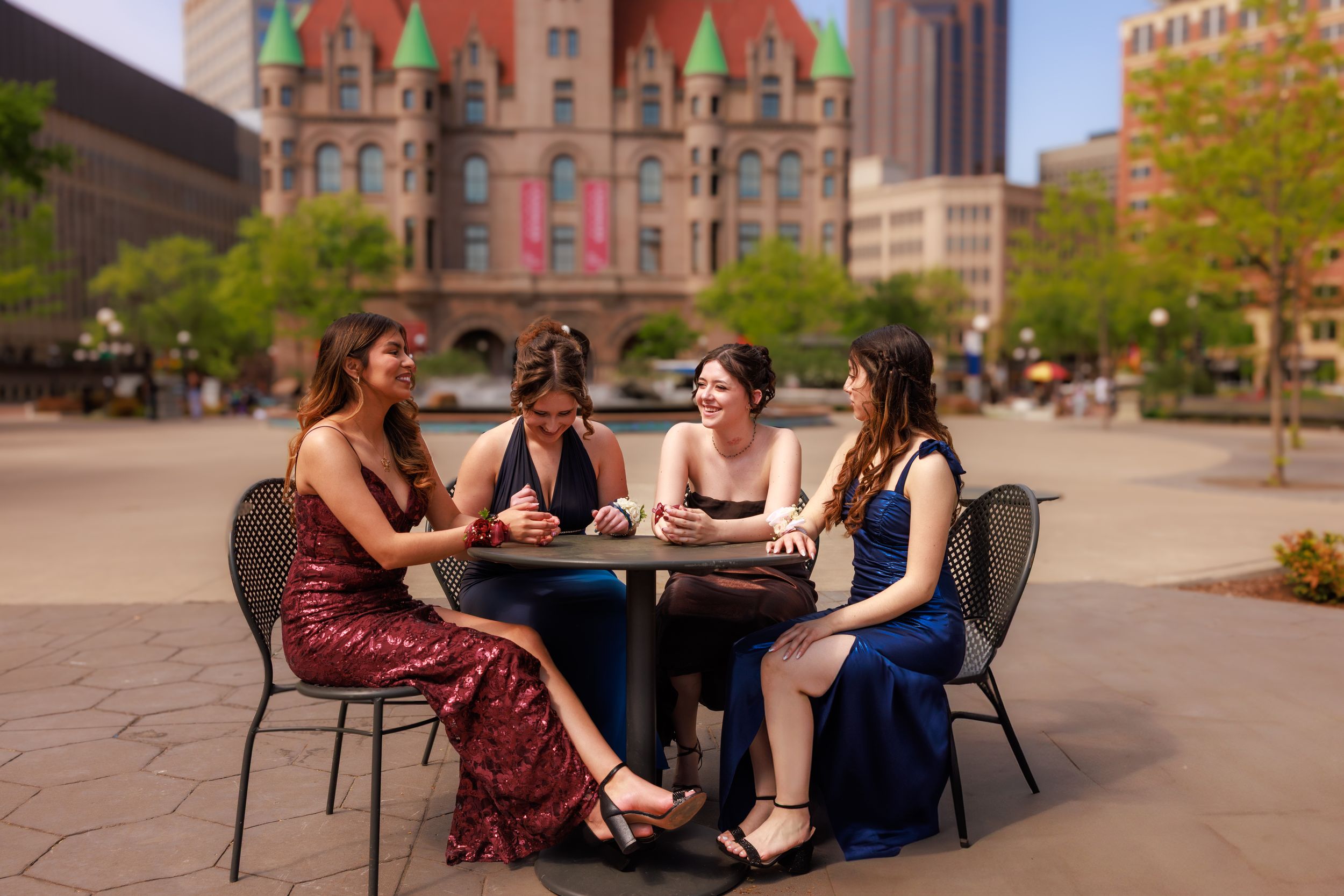 Four young women in formal dresses sit together at an outdoor table in a city plaza on a sunny day.