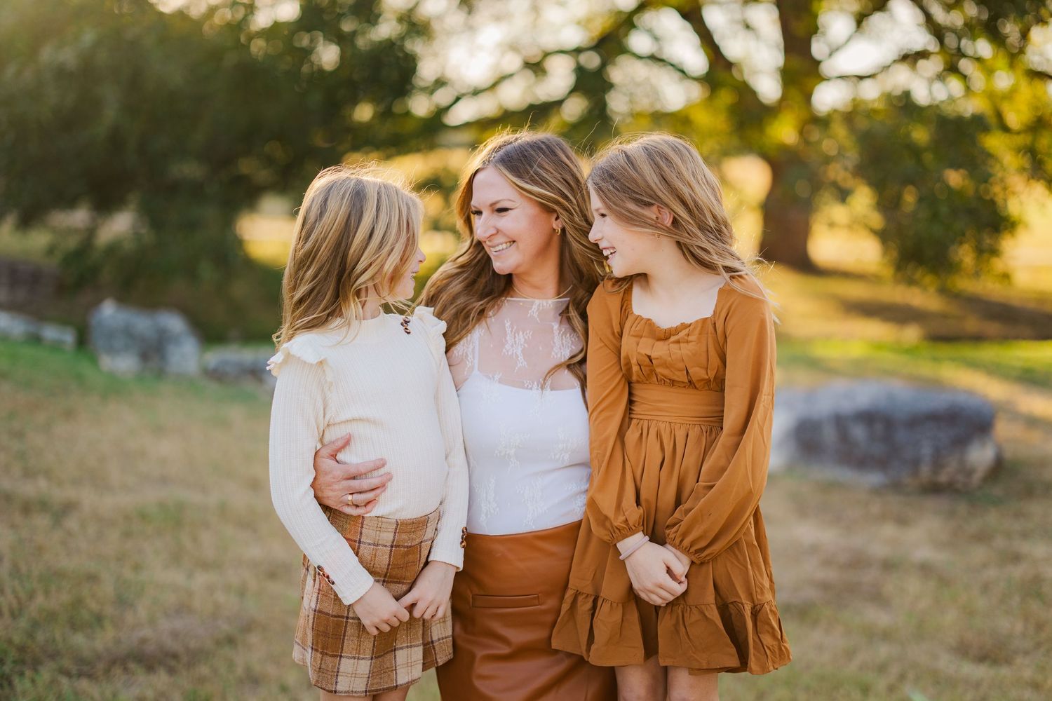 A mother smiles warmly with her two blonde daughters in a sunlit field, all wearing coordinating brown and cream outfits.