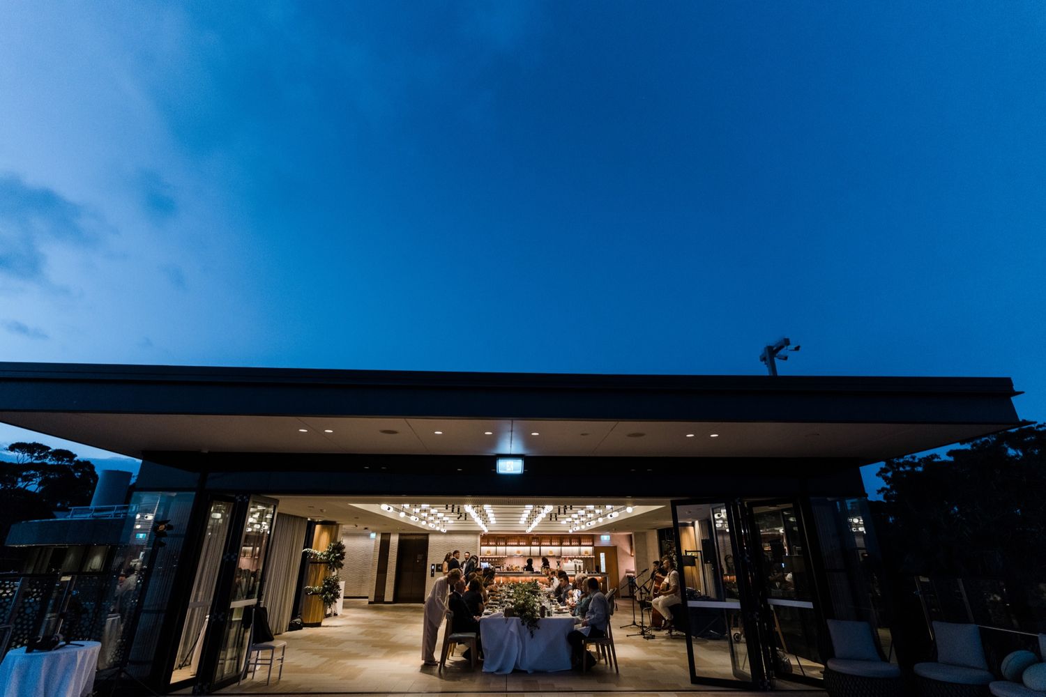 Elegant venue exterior at dusk with warm interior lighting and guests gathered under a modern open-air pavilion.