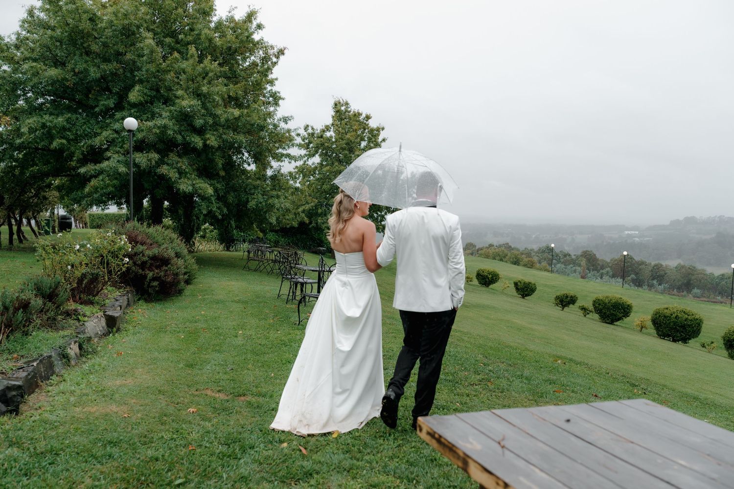 Bride and groom share umbrella on lush green hillside during rainy outdoor wedding ceremony. Borrodell Vineyard Orange. Orange wedding