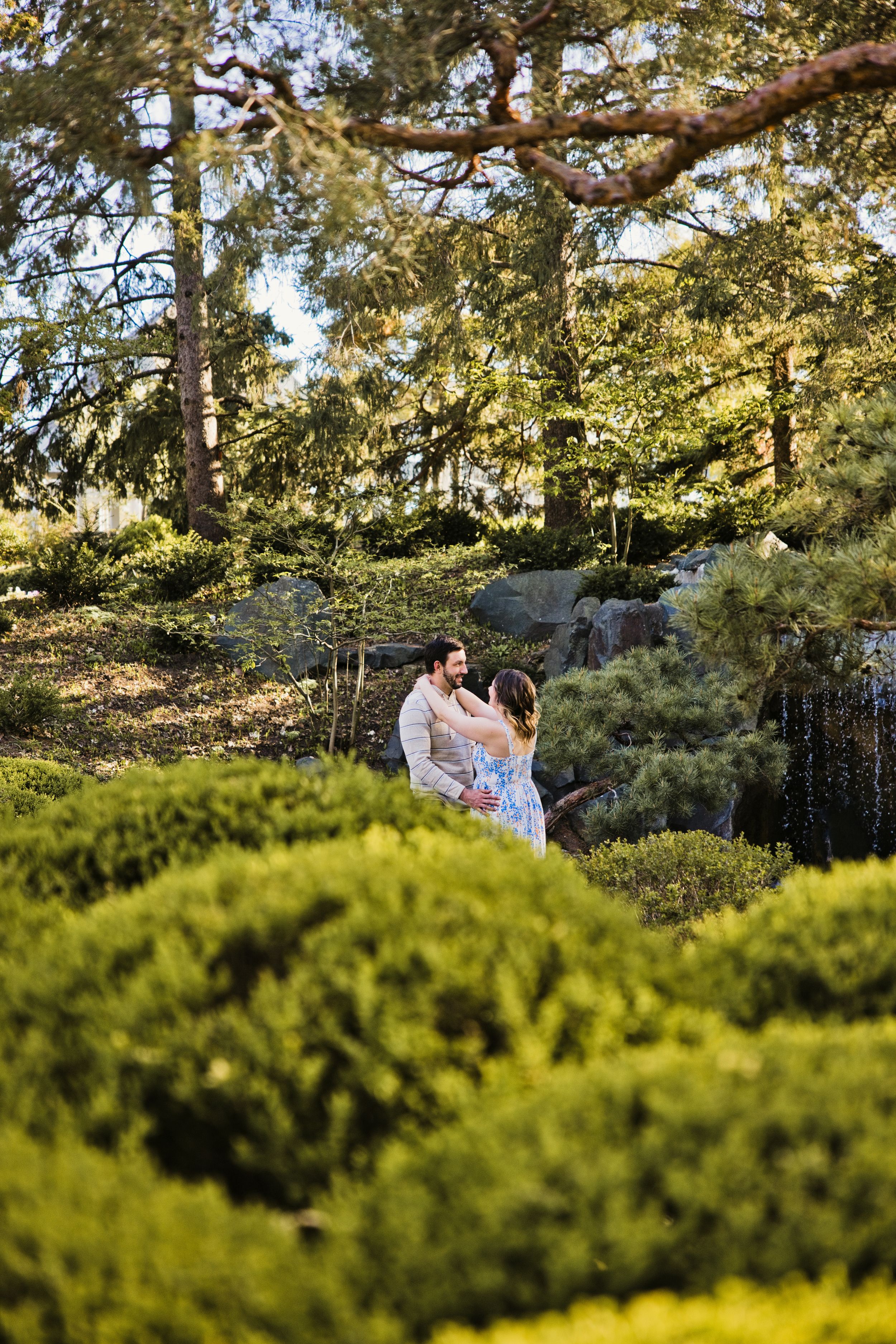 Couple embracing in a lush green garden surrounded by trimmed hedges, pine trees, and rocky waterfall backdrop.