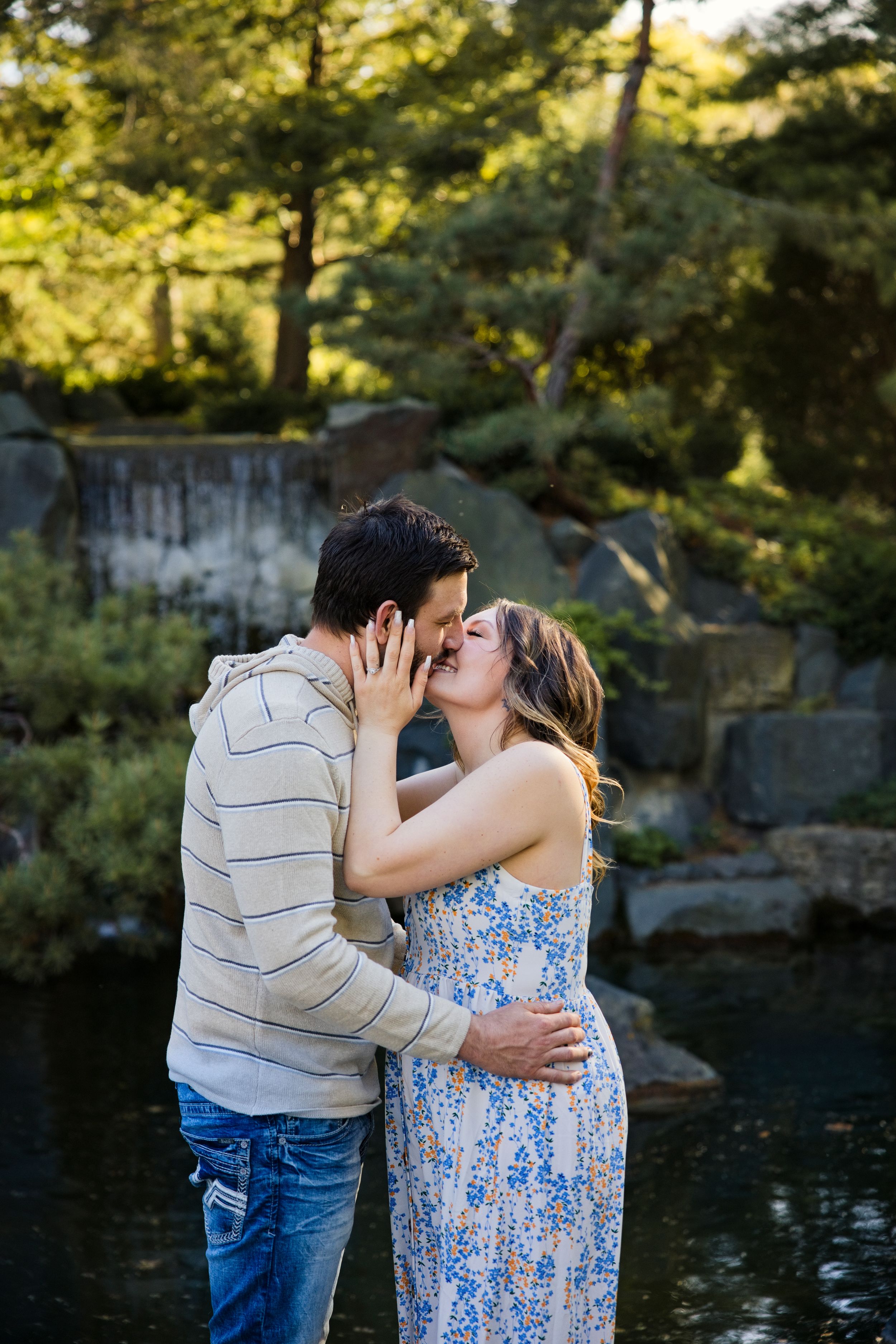 Couple sharing a romantic kiss by a serene garden pond, woman in floral dress, man in striped hoodie and jeans.