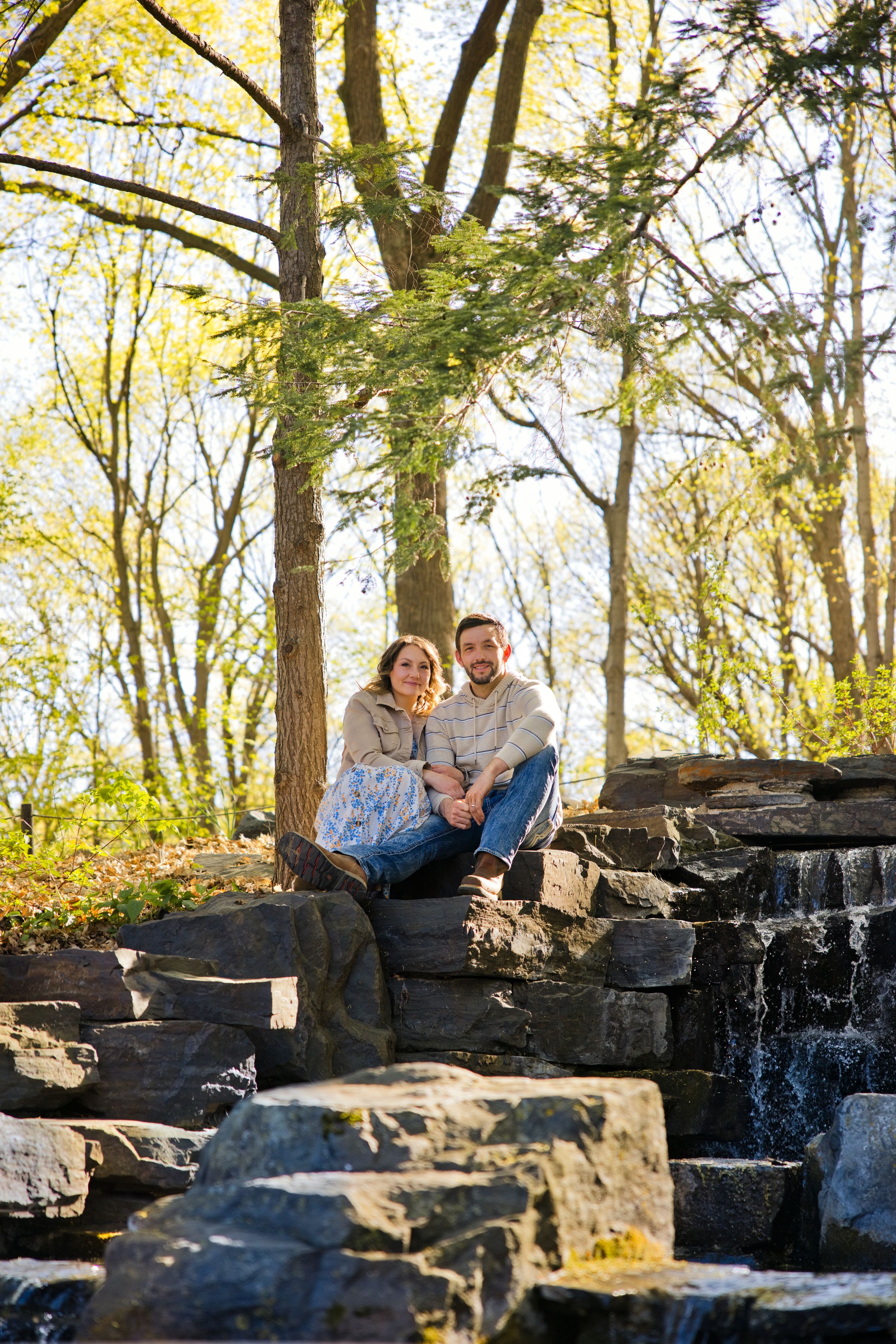 Couple sitting together on stone steps near a waterfall surrounded by trees in a park during spring.