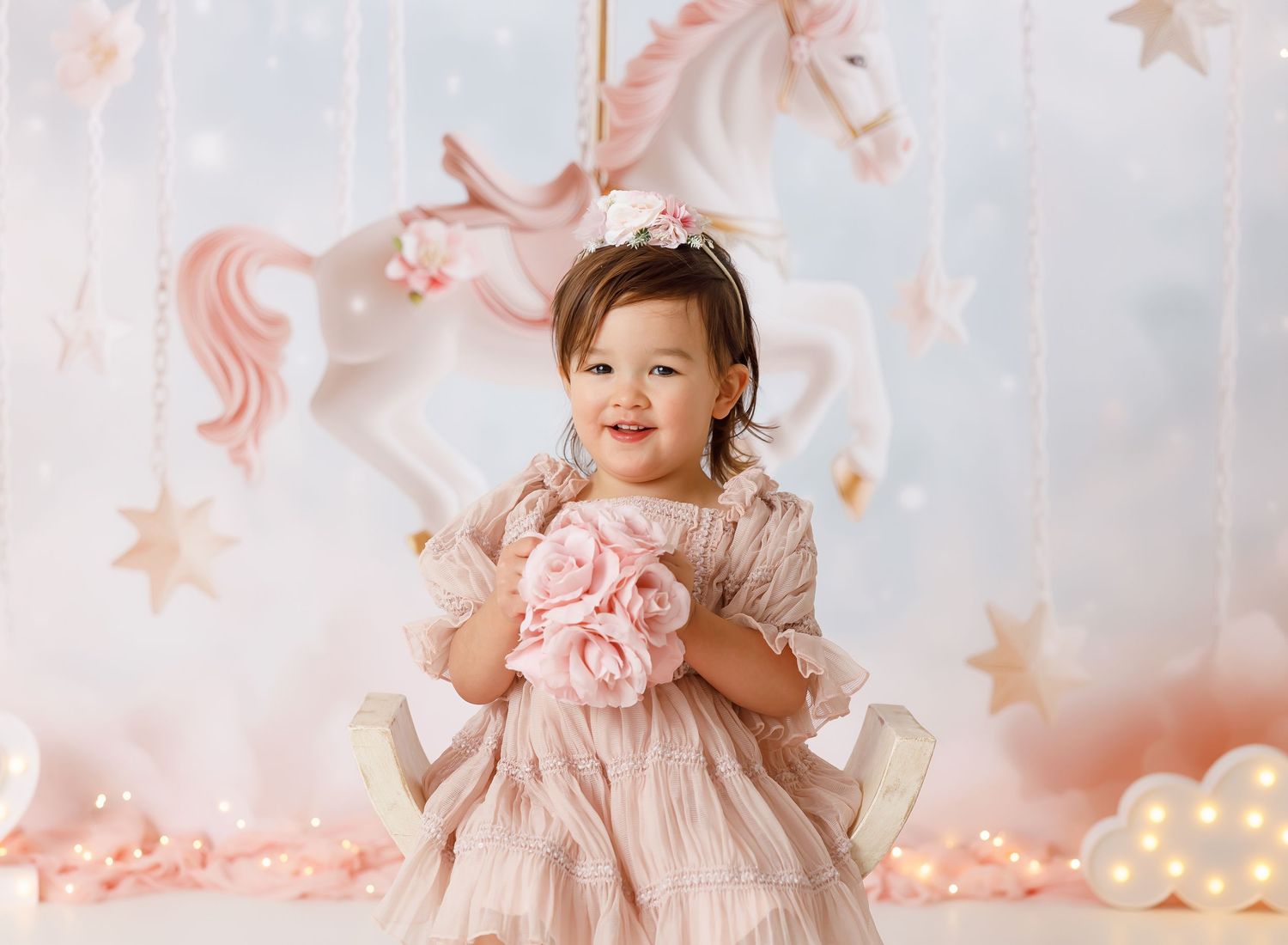 Toddler girl in pink dress holding roses, smiling in front of carousel unicorn backdrop with stars.