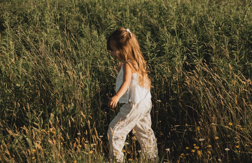 A young girl in floral pants walks through a tall green meadow field in warm golden sunlight.