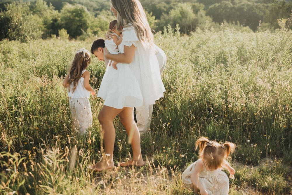A mother and three young daughters in white dresses play in a sunny green meadow during a family photo session.
