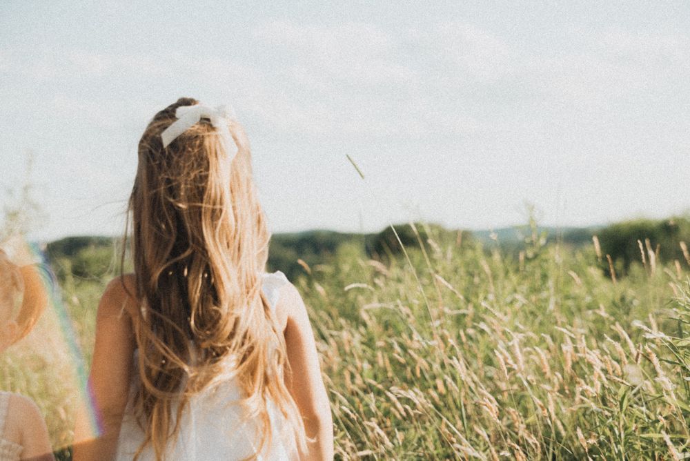 Young girl with long blonde hair standing in a sunny meadow with tall grass, viewed from behind.