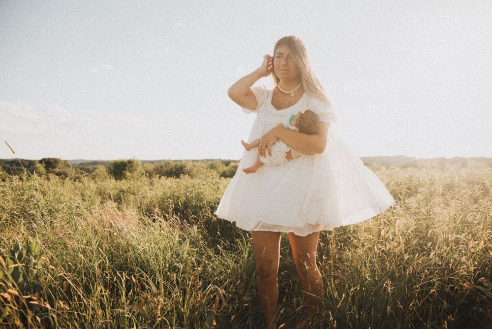 A woman in a white dress breastfeeds her baby in a sunlit field of tall green grass.
