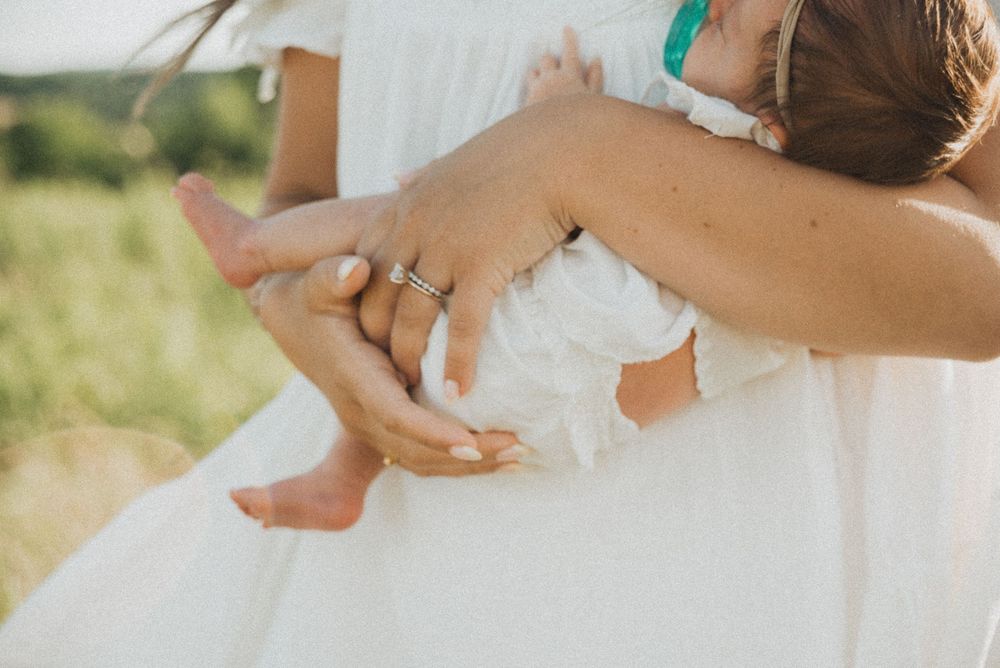 A mother in a white dress holds her barefoot child outdoors in a sunny green field, wearing a diamond ring.