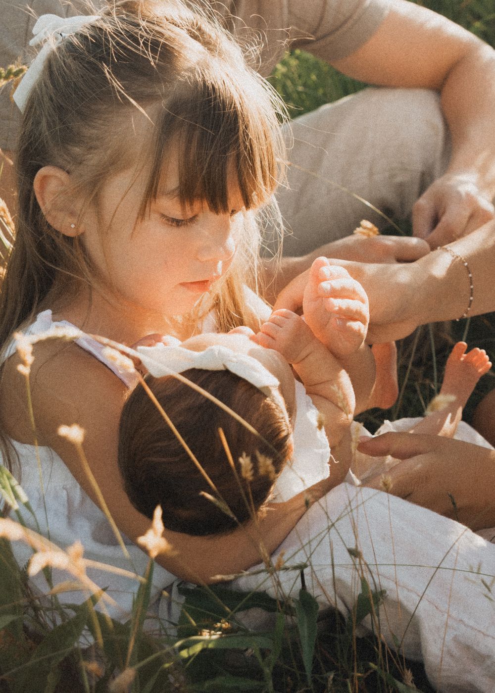 A young girl sits outdoors in golden sunlight, gently holding a newborn baby surrounded by wildflowers.