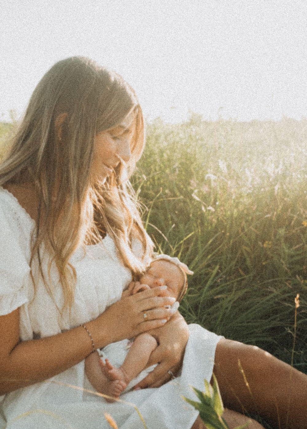 A young mother in a white dress sits in a sunlit field, tenderly cradling her newborn baby outdoors.