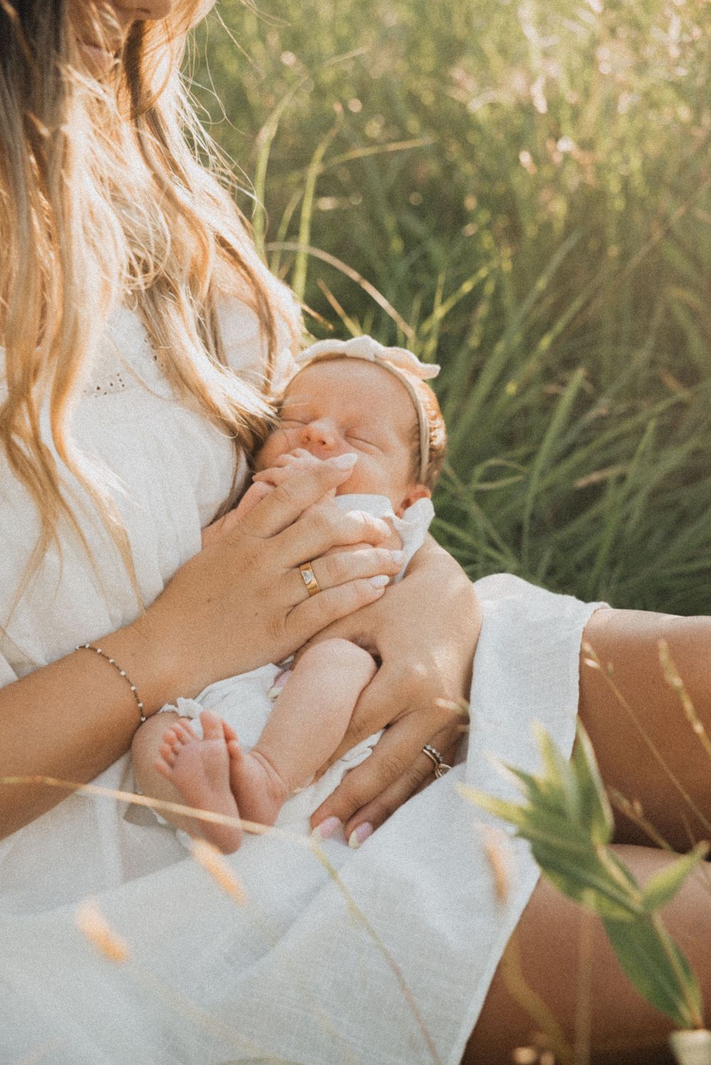Mother in white dress bottle feeding newborn baby outdoors in golden sunlight among tall green grass.