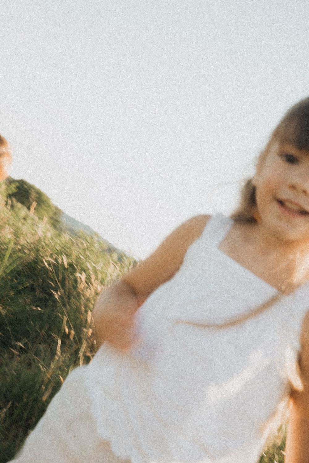 Two young girls in white dresses running through a sunny green meadow, laughing and playing outdoors.